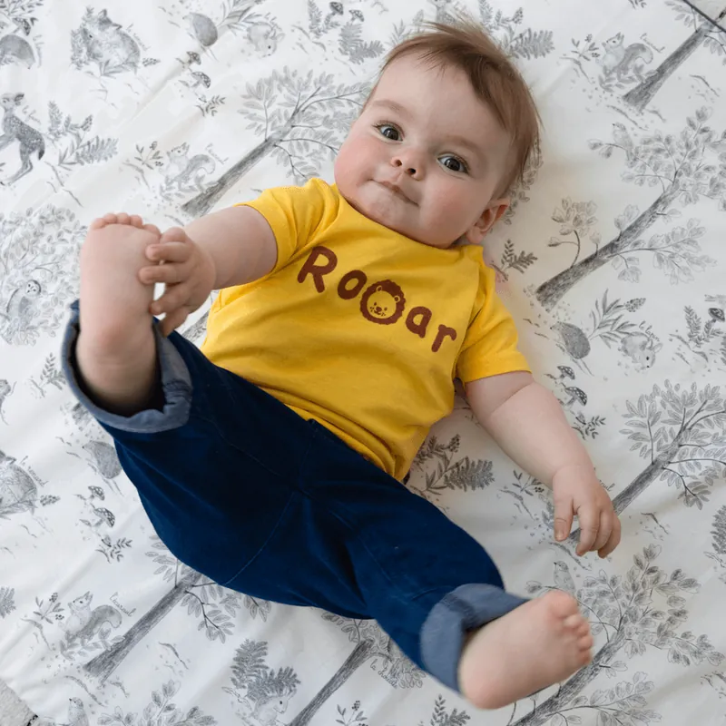 Un bébé en tee-shirt jaune « Rooar » et jean, allongé sur le dos, attrape son pied sur un tapis d'éveil gris à motif d'animaux de la forêt