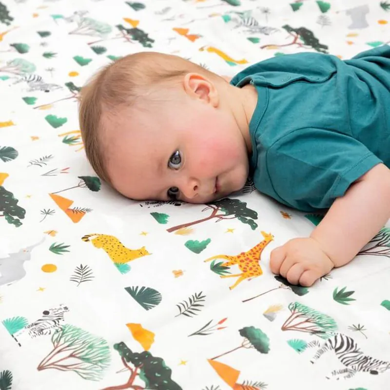 Un bébé en tee-shirt bleu canard, la joue posée sur un tapis d'éveil blanc imprimé savane, girafe et zèbres