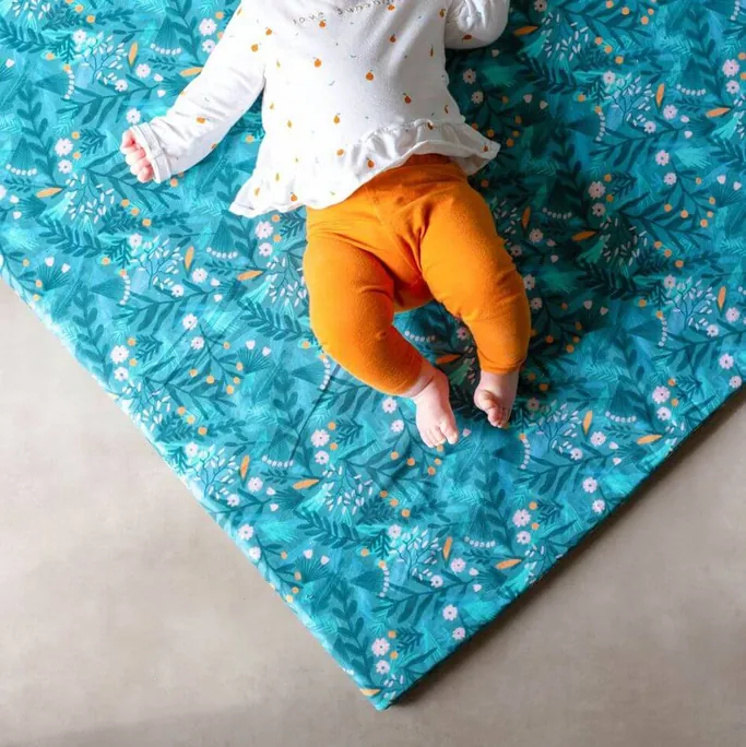 Un bébé en haut blanc et pantalon orange, sur le ventre, sur un tapis d'éveil bleu canard à motif de feuillage fleuri posé sur un sol gris