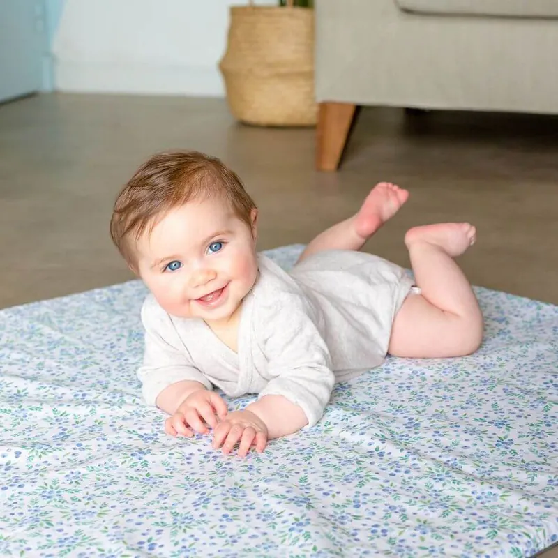 Un bébé souriant en body blanc, sur le ventre, sur un tapis d'éveil blanc à petites fleurs bleues, dans un salon