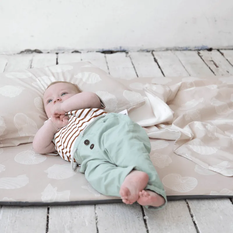 Bébé allongé sur le dos sur un tapis beige à motifs de feuilles, la main à la bouche, la tête sur un oreiller assorti