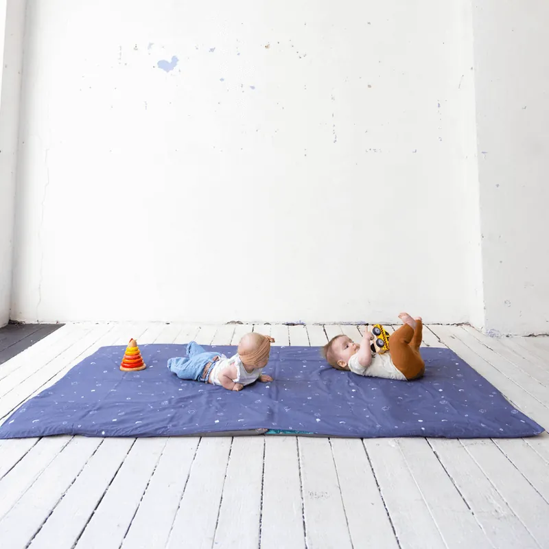 Deux bébés allongés sur un grand tapis d'éveil bleu nuit étoilé, dans une pièce blanche au parquet peint, un anneau à empiler posé à côté