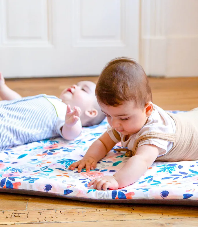 Deux bébés sur un tapis d'éveil à fleurs posé sur un parquet : l'un allongé sur le dos, l'autre sur le ventre qui tend la main