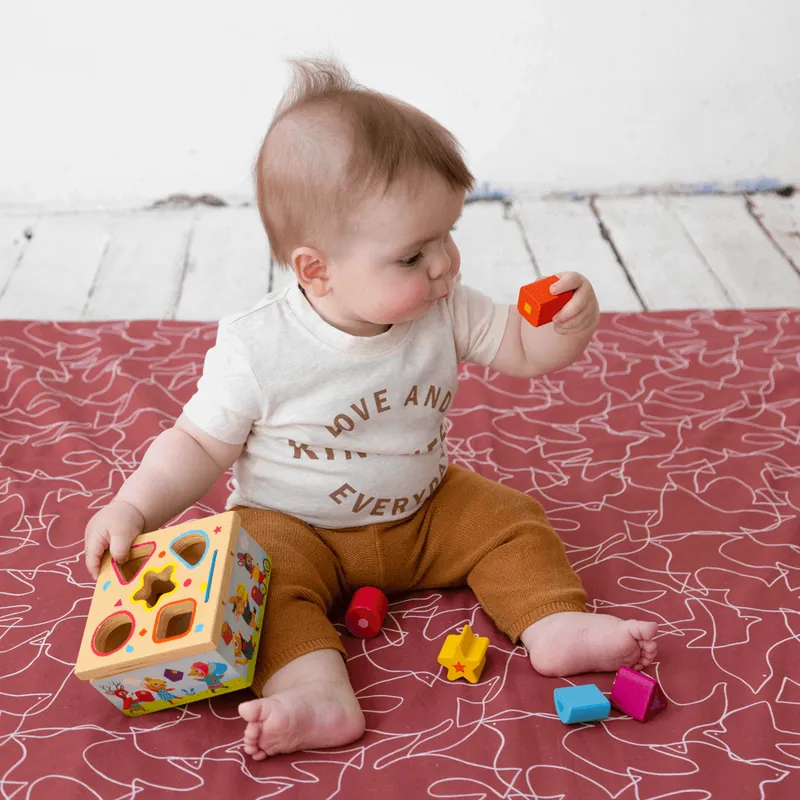 Bébé assis sur un tapis rouge à motifs blancs, qui joue avec une boîte à formes en bois entourée de formes colorées