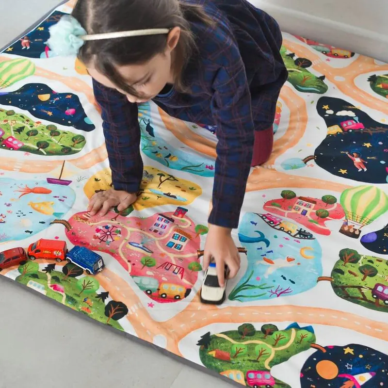 Petite fille à genoux sur un grand tapis illustré de routes, de planètes et de montgolfières, qui fait rouler une petite voiture