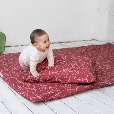 Bébé souriant à quatre pattes sur un tapis d'éveil rouge à motifs blancs, près d'un oreiller assorti