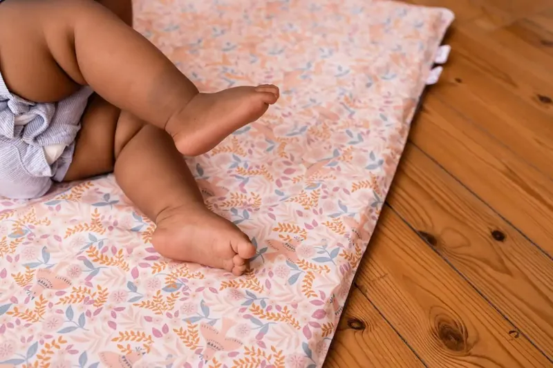 Jambes et pieds d'un bébé sur un tapis d'éveil rose à motifs de fleurs, posé sur un parquet