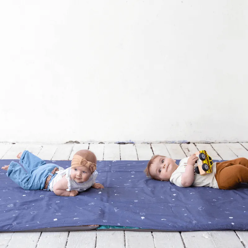 Deux bébés allongés sur un grand tapis bleu nuit étoilé, l'un sur le ventre, l'autre sur le dos avec une petite voiture jaune