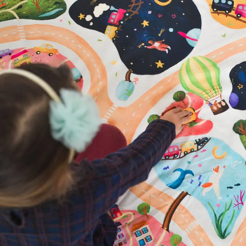 Petite fille avec un bandeau à pompon, vue de dos, qui joue sur un tapis illustré de routes, de planètes et de montgolfières