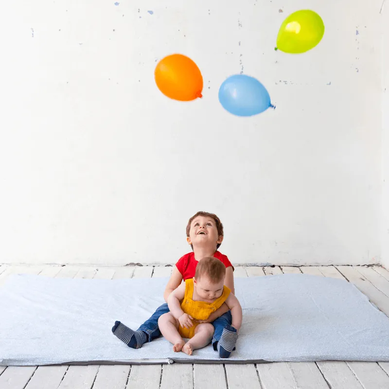 Un petit garçon en tee-shirt rouge rit, assis sur un grand tapis bleu clair, un bébé sur les genoux, sous trois ballons de baudruche