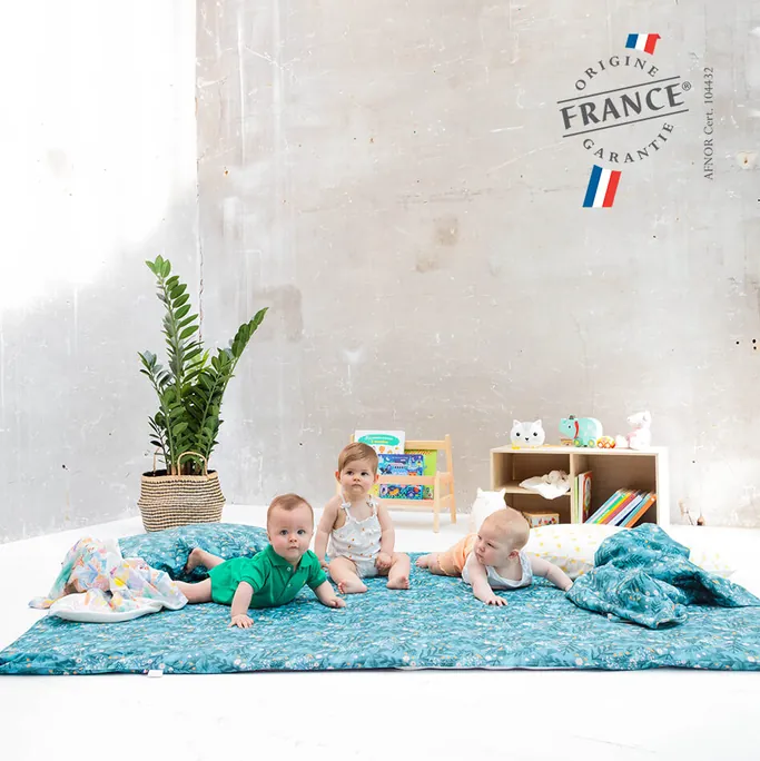 Trois bébés jouent sur une grande housse turquoise étalée au sol, devant une étagère à jouets et une plante verte, logo Origine France Garantie en haut à droite