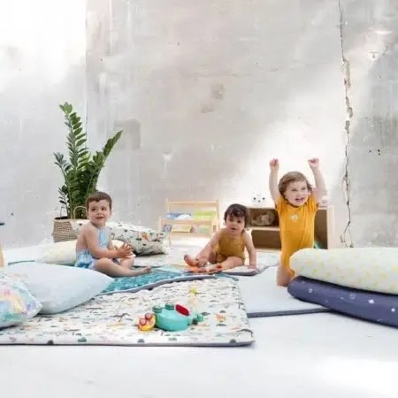 Trois jeunes enfants jouent sur des tapis d'éveil dépliés au sol, dans une pièce aux murs de béton clair avec une plante verte