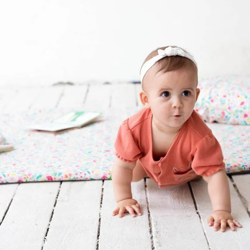 Une petite fille au bandeau blanc, en barboteuse corail, avance à quatre pattes devant un tapis d'éveil fleuri posé sur un plancher blanc