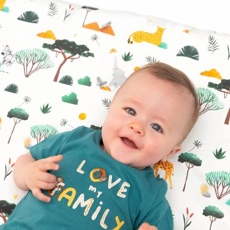 Un bébé souriant en t-shirt vert « Love my family », allongé sur un tapis d'éveil imprimé savane avec arbres, girafe et guépard