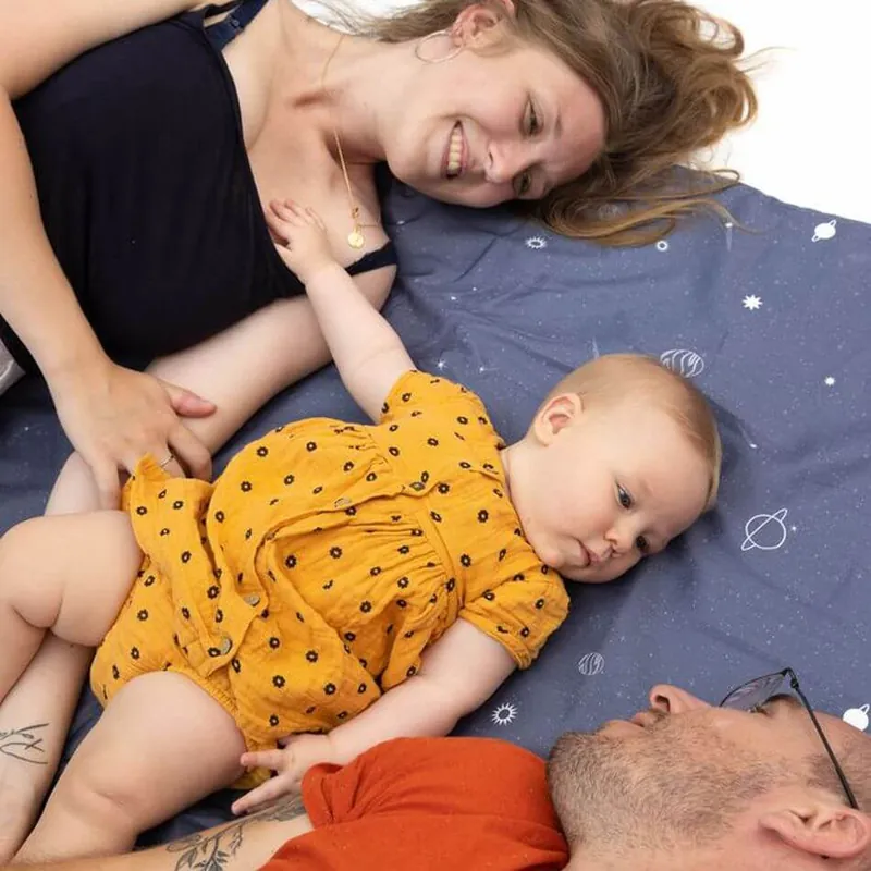 Une maman et un papa allongés sur un tapis d'éveil bleu nuit étoilé, autour de leur bébé en barboteuse moutarde