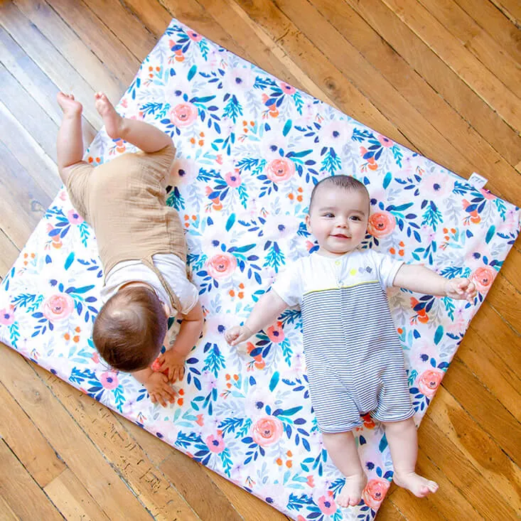 Deux bébés sur un tapis d'éveil fleuri posé sur un parquet, l'un sur le ventre, l'autre souriant sur le dos
