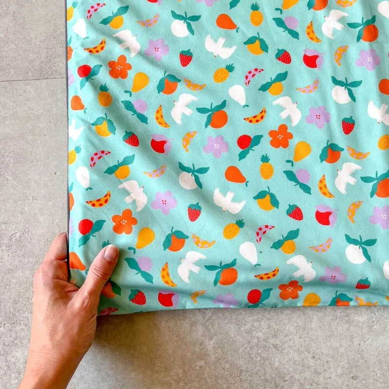 Une main soulève le coin d'un tapis d'éveil turquoise imprimé de fruits et d'oiseaux, posé sur un sol gris