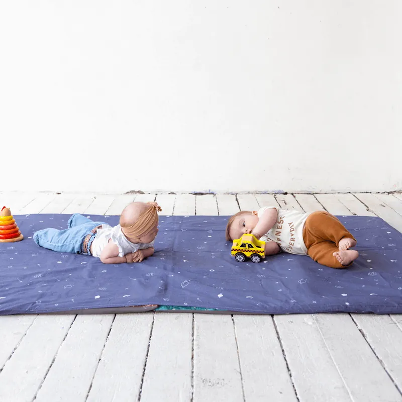 Deux bébés jouent au sol sur un tapis d'éveil bleu nuit étoilé, l'un avec une petite voiture jaune, l'autre à côté d'une pyramide à anneaux