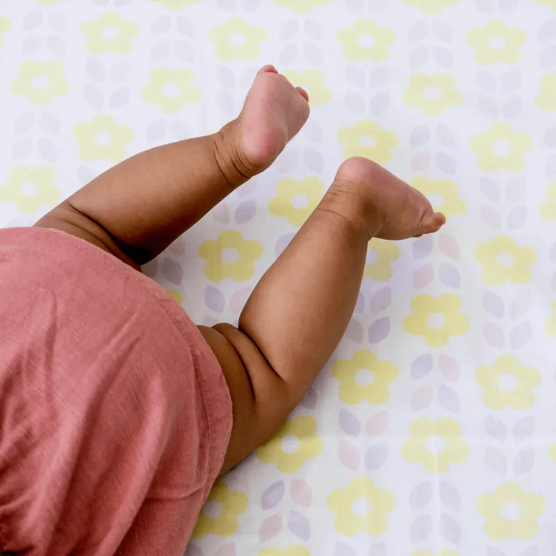 Gros plan sur les jambes d'un bébé en body rose, allongé sur un tapis d'éveil à fleurs jaunes et lilas