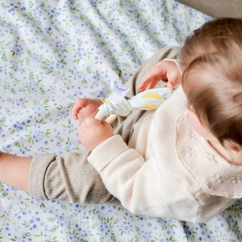 Un bébé en gilet écru, assis sur un tapis à petites fleurs bleues, tient un anneau de dentition en bois garni de tissu