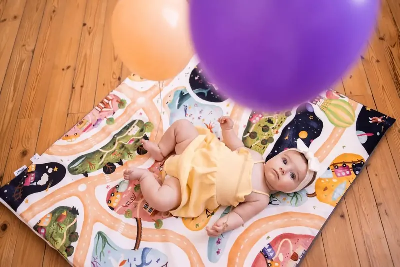 Un bébé en barboteuse jaune, allongé sur un tapis d'éveil imprimé de routes et de montgolfières posé sur un parquet, sous des ballons violet et pêche