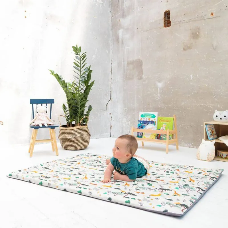 Un bébé en t-shirt vert, à plat ventre, relève la tête sur un tapis d'éveil blanc à petits motifs colorés, dans une pièce aux murs de béton clair