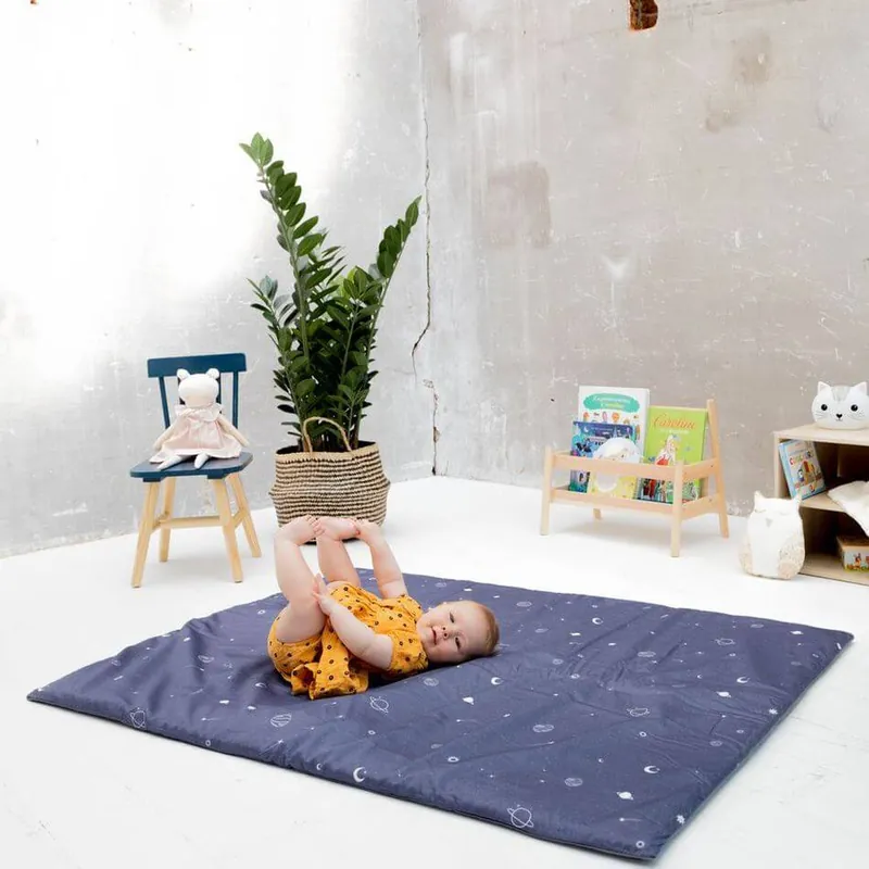 Un bébé en barboteuse moutarde, allongé sur le dos, attrape ses pieds sur un tapis d'éveil bleu nuit étoilé, devant une petite chaise et une plante verte