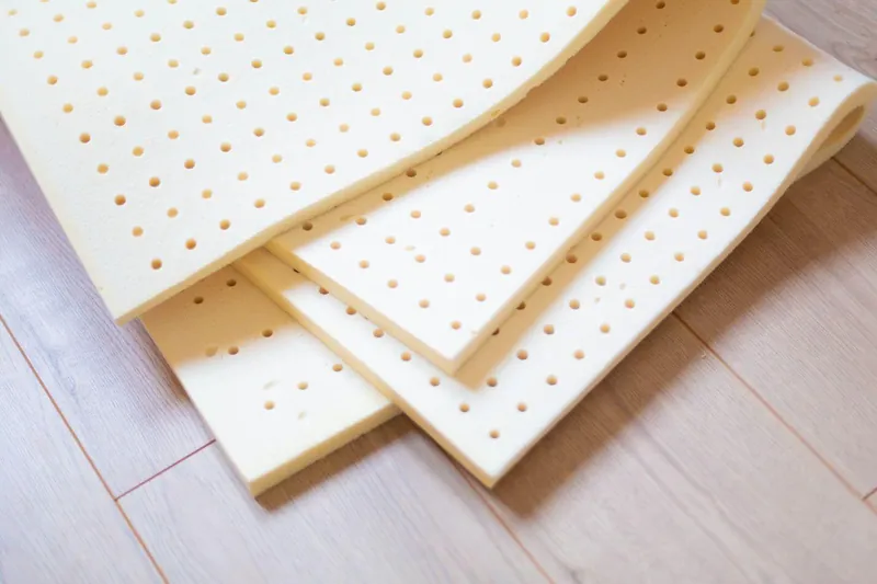 Trois plaques de mousse en latex naturel perforées, couleur crème, posées en éventail sur un parquet clair