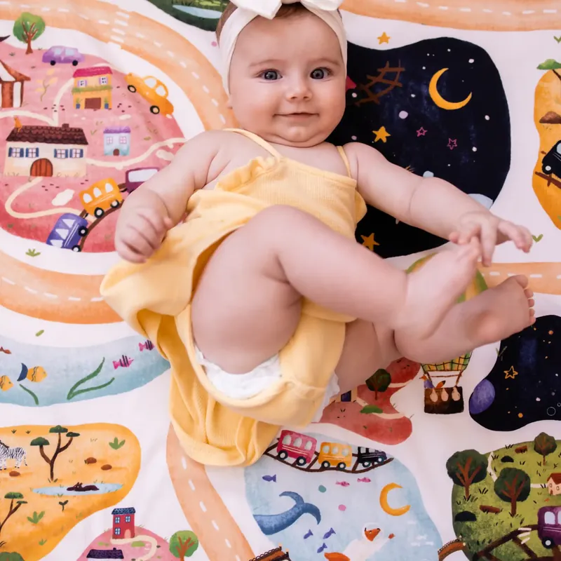 Un bébé en robe jaune, allongé sur le dos, attrape son pied sur un tapis d'éveil imprimé de routes, de maisons et d'un ciel étoilé