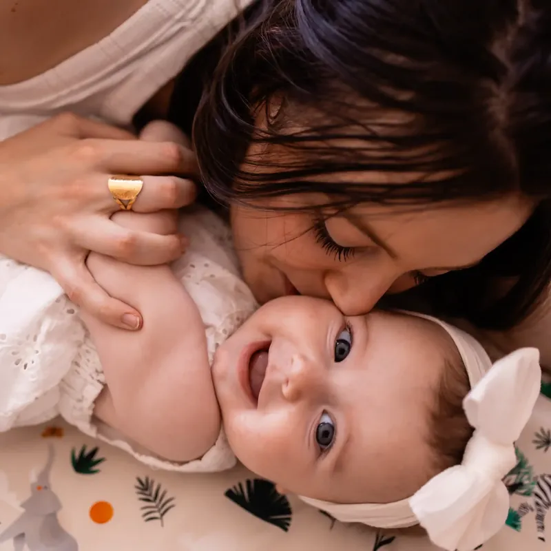 Une maman embrasse la joue de son bébé souriant, coiffé d'un gros noeud blanc, allongé sur un tapis d'éveil imprimé de feuillages