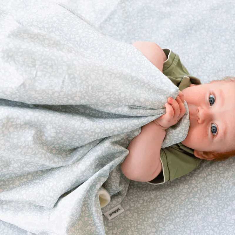 Un bébé en t-shirt kaki, allongé sur un tapis d'éveil gris clair à petits motifs, tire le bord de la housse vers sa bouche