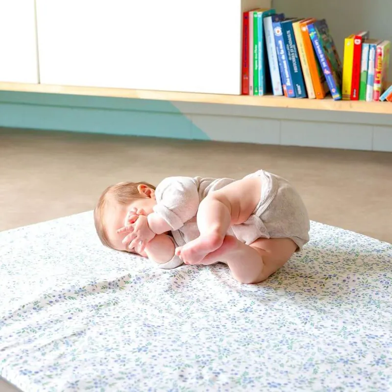 Un bébé en body blanc, recroquevillé sur un tapis d'éveil à petites fleurs bleues, devant une étagère de livres colorés