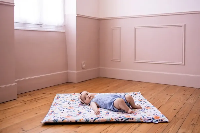 Un bébé en barboteuse bleue allongé sur le dos sur un tapis d'éveil fleuri, au milieu du parquet d'une chambre aux murs roses