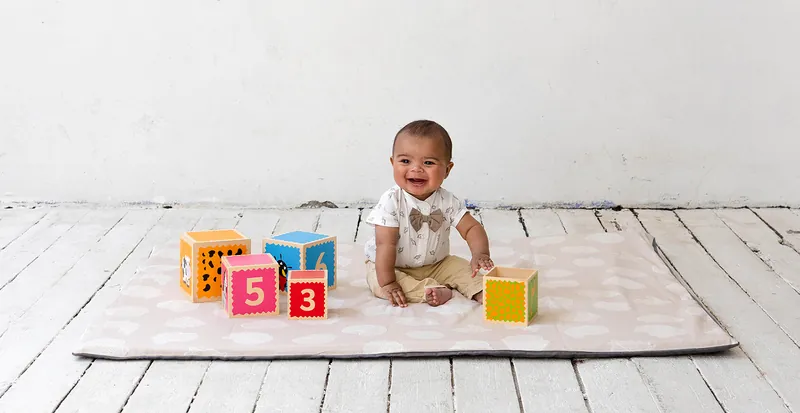 Un bébé souriant, assis sur un tapis d'éveil beige, joue avec des cubes à chiffres colorés sur un plancher blanc