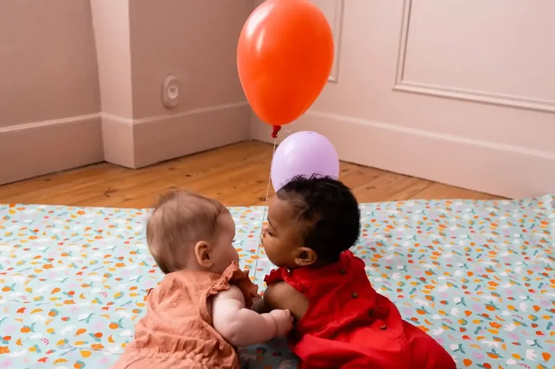 Deux bébés, l'un en rose, l'autre en robe rouge, jouent face à face sur un tapis d'éveil moucheté, sous deux ballons rouge et mauve