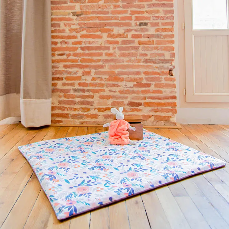 Un doudou lapin rose posé sur un tapis d'éveil à fleurs bleues et roses, déplié sur un parquet devant un mur de briques