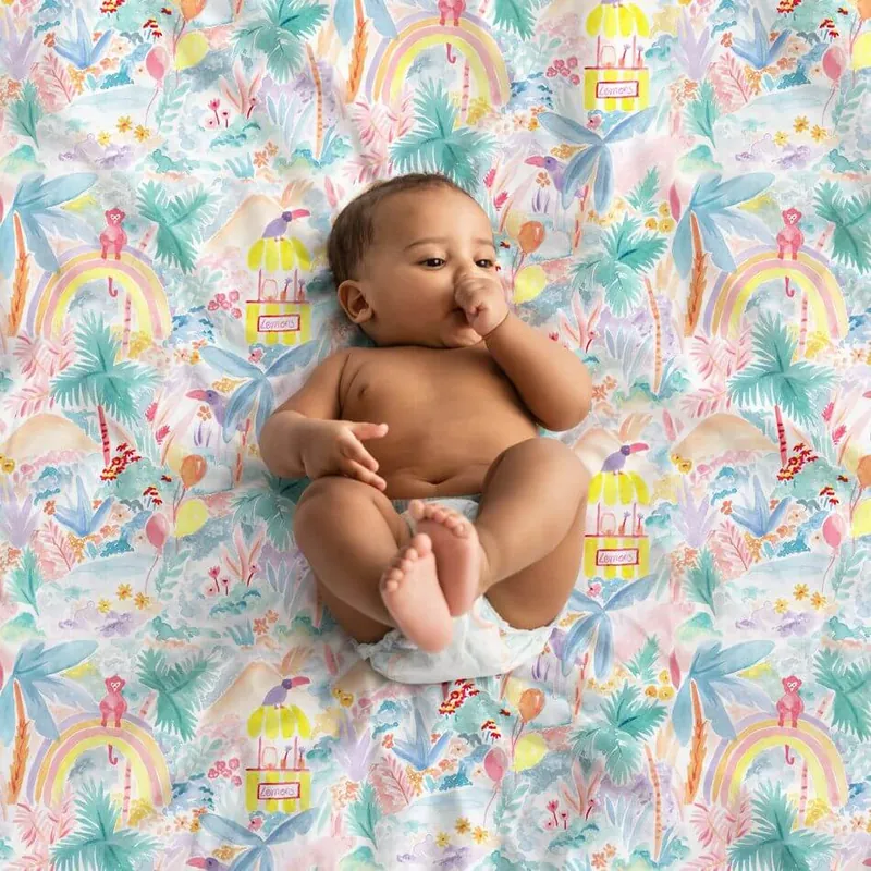 Un bébé en couche, allongé sur le dos, suce son pouce sur un tapis d'éveil pastel imprimé de jungle, d'arcs-en-ciel et de palmiers
