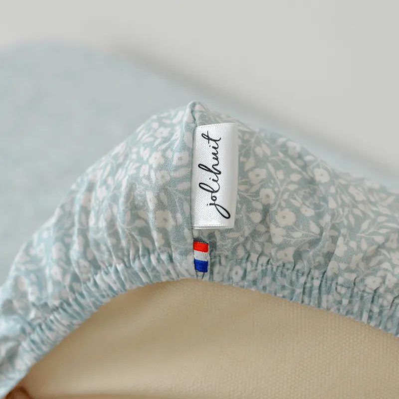 Gros plan sur le coin d'une housse bleu clair à fleurs blanches bordée d'un élastique, avec l'étiquette Jolihuit et un petit drapeau français, sur une mousse de latex crème