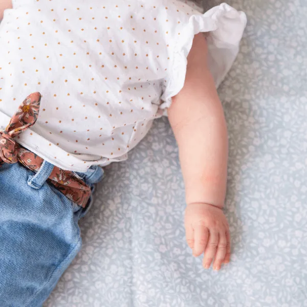Un bébé en blouse blanche à pois et jean à ceinture fleurie, allongé sur un tapis d'éveil bleu clair à petites fleurs