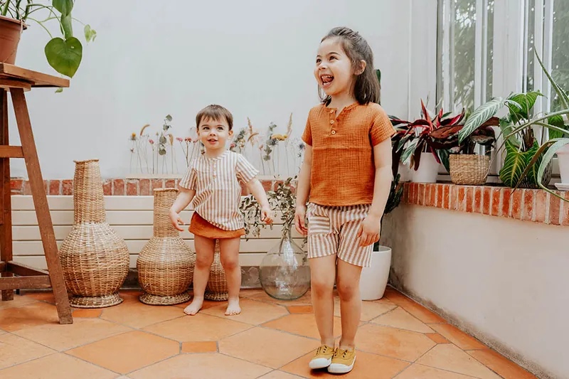 Deux enfants rieurs, en ensemble rayé et en tenue orange, debout sur un carrelage de terre cuite au milieu de plantes vertes