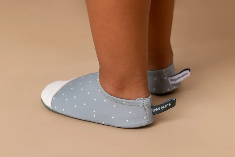 Pieds d'enfant chaussés de chaussons souples gris à pois blancs, sur fond beige