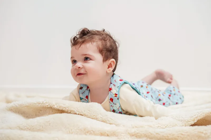 Un petit garçon aux cheveux bouclés, en gilet imprimé bleu, allongé sur le ventre sur une couverture en laine bouclée