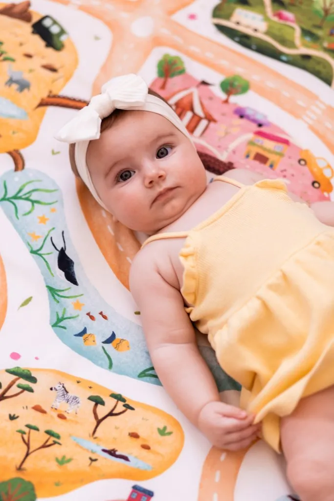 Un bébé en barboteuse jaune, un bandeau noué dans les cheveux, allongé sur un tapis imprimé d'une carte illustrée