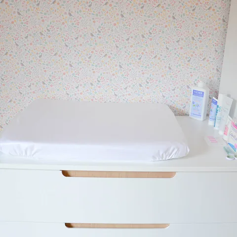 Matelas à langer en housse imperméable blanche sur la commode, flacons de soin et papier peint fleuri