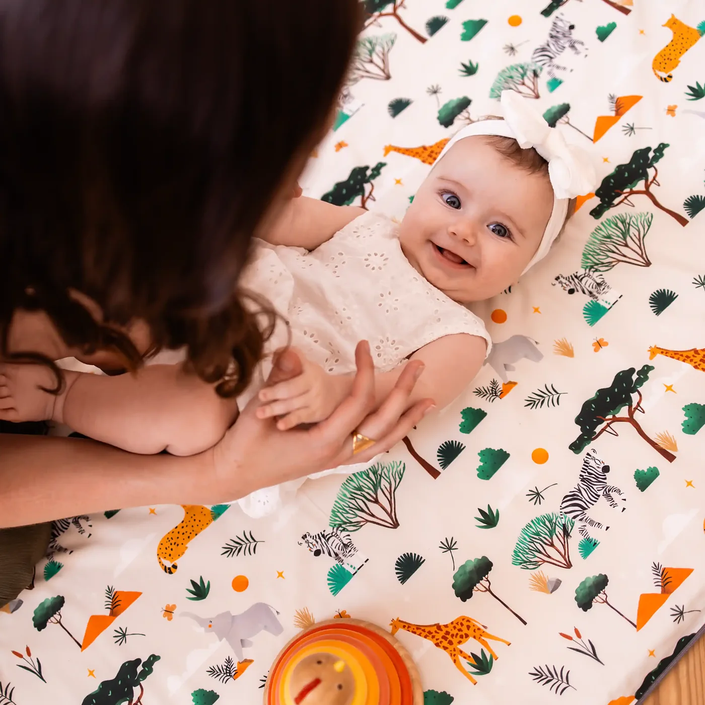 Le bébé sourit dans les bras de sa mère, sur le tapis imprimé savane, à côté d’un jouet en bois.